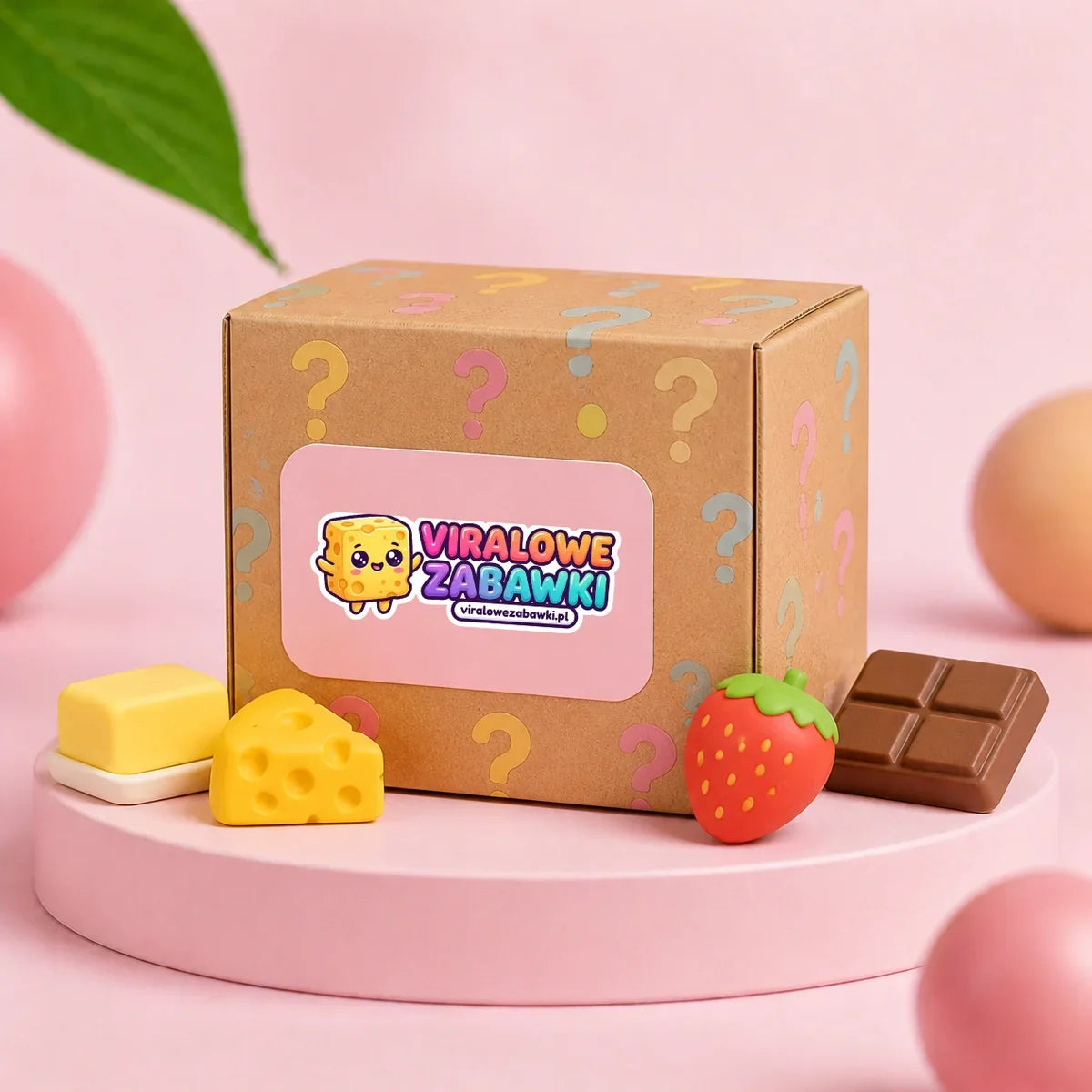 Średnie kartonowe pudełko Mystery Box z logo sklepu i znakami zapytania, obok przykładowe gniotki squishy: masło, ser, truskawka i czekolada, na różowym podeście