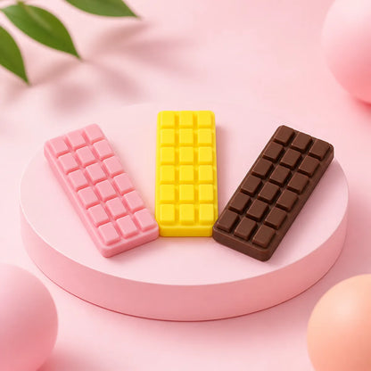 Trzy gniotki squishy w kształcie tabliczek czekolady: różowa, żółta i ciemnobrązowa na okrągłym różowym podeście